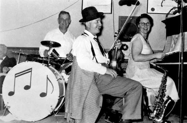 Photo en noir et blanc de trois musiciens adultes assis et souriants ; deux hommes et une femme, portant des instruments.