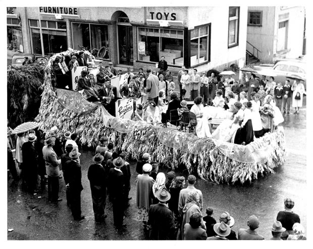 Photo en noir et blanc montrant une rue pavée bordée de spectateurs observant une remorque décorée comme un char de parade, dont les côtés sont ornés de branches de cèdre. À l'avant de la remorque, un groupe musical d'environ 15 personnes est dirigé par un homme debout face à eux, tournant le dos à environ 8 personnes assises à l'arrière du char.