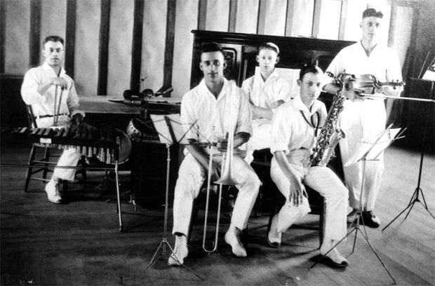 Photo en noir et blanc de cinq jeunes musiciens masculins aux cheveux courts, portant des instruments et vêtus de manière identique : chemises blanches à col ouvert, pantalons blancs et chaussures blanches.