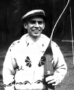 Photo en noir et blanc d'un homme d'âge moyen au visage rude, portant une casquette et un épais cardigan en laine, tenant un arc en bois dans sa main gauche.
