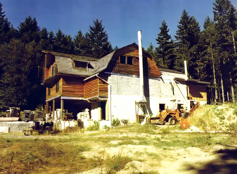 Photo en couleur d’un vaste bâtiment de style grange, à trois étages et en forme de croix, dont la partie centrale est construite en béton et en blocs de béton, tandis que les extrémités et les étages supérieurs sont en bois.