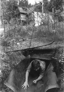 Photo en noir et blanc d’un homme rampant hors d’un tunnel souterrain. À environ 100 mètres derrière lui, une grange à trois étages se dresse en arrière-plan.
