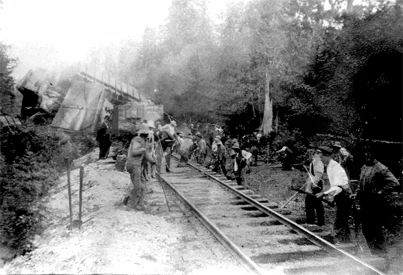 Photo en noir et blanc d'une voie ferrée au premier plan le long de laquelle travaillent des groupes d'hommes, dont de nombreux Chinois. À l'arrière-plan se trouve l'épave de nombreux wagons de charbon en bois entassés ensemble en une pile désordonnée.
