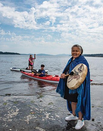 Photographie en couleur d'une femme Coast Salish avec de longs cheveux gris, portant une cape azur fermée au cou sur une robe noire mi-genoux et des baskets blanches. Elle tient une baguette et un tambour en peau rond décoré d'animaux stylisés Coast Salish. Elle se tient sur une rive rocheuse. À l'arrière, une personne est assise dans un kayak et une autre est agenouillée sur une planche à pagaie. Des îles boisées forment l'horizon.