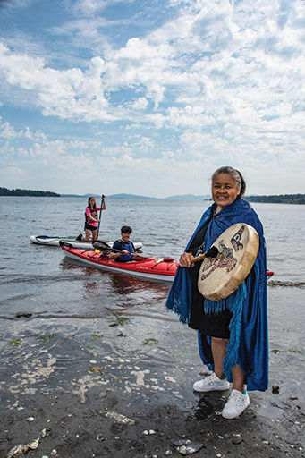 Photographie en couleur d'une femme Coast Salish avec de longs cheveux gris, portant une cape azur fermée au cou sur une robe noire mi-genoux et des baskets blanches. Elle tient une baguette et un tambour en peau rond décoré d'animaux stylisés Coast Salish. Elle se tient sur une rive rocheuse. À l'arrière, une personne est assise dans un kayak et une autre est agenouillée sur une planche à pagaie. Des îles boisées forment l'horizon.