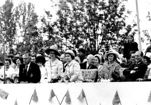 Photographie en noir et blanc de dignitaires assis en plusieurs rangées, regardant des festivités hors du cadre de la photo.
