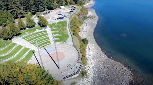 Photographie aérienne en couleur d'une scène circulaire en ciment et briques située au bord de l'eau. Deux grands poteaux en bois se dressent légèrement décentrés du côté maritime de la scène. Côté terre, face à la scène, une aire semi-circulaire en pente est aménagée en gradins recouverts de gazon.