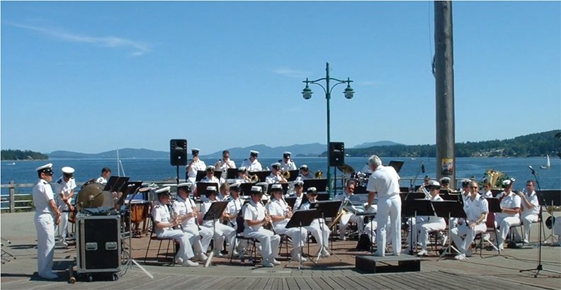 Photographie en couleur d’une fanfare d’une trentaine de musiciens en uniformes blancs, jouant sur une scène en plein air, avec en arrière-plan une vue sur la mer et des îles boisées, le tout sous un ciel bleu presque sans nuages.