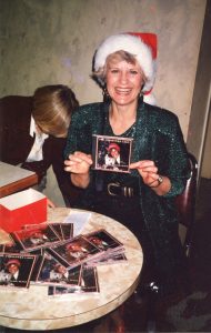 Photo en couleur d'une femme blonde, souriante, vêtue d'une robe vert foncé pailletée et d'un chapeau de Noël rouge et blanc, assise devant une table ronde remplie de pochettes de disques compacts, tenant une pochette de CD avec sa photo et le titre The Christmas Lady.
