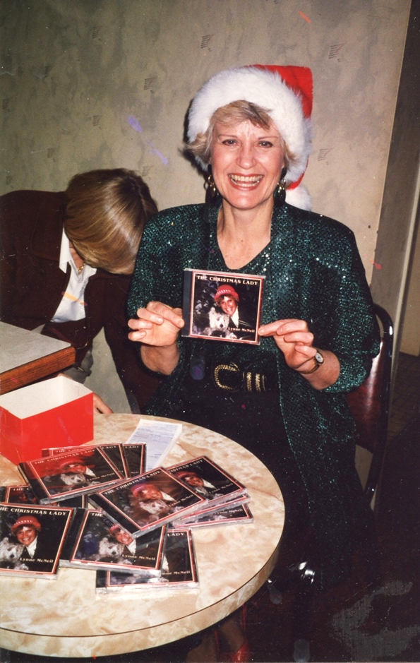 Photo en couleur d'une femme blonde, souriante, vêtue d'une robe vert foncé pailletée et d'un chapeau de Noël rouge et blanc, assise devant une table ronde remplie de pochettes de disques compacts, tenant une pochette de CD avec sa photo et le titre The Christmas Lady.