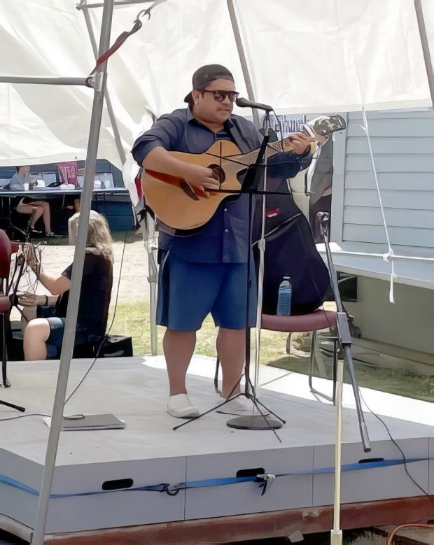 Photo en couleur d'un musicien Salish de la côte sur scène, devant un microphone, jouant de la guitare acoustique électrifiée et chantant.