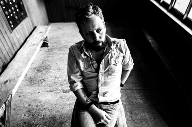 Photo en noir et blanc d'un homme barbu assis, les cheveux coupés au-dessus des oreilles et coiffés à gauche, portant une chemise de style Wrangler, les manches longues retroussées jusqu'aux coudes, et un jean en denim.