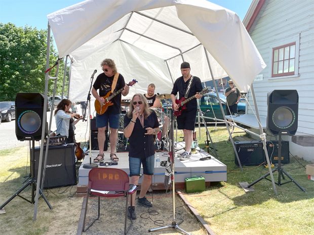 Photo en couleur d'un groupe de rock de cinq musiciens jouant sur une petite scène extérieure, à l'abri du soleil sous une tente blanche.