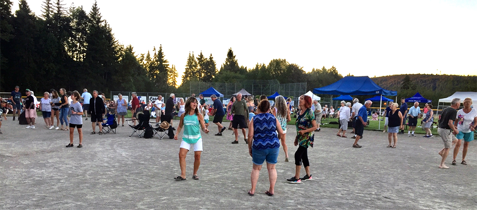 Photo en couleur d'un grand groupe, plusieurs portant des t-shirts et des shorts, sur un terrain de sport lors d'une belle soirée d'été, dansant et écoutant de la musique.