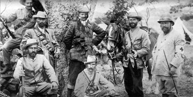 Photographie en noir et blanc montrant sept hommes posant avec des fusils, prêts au combat. Un cheval et une tente apparaissent en arrière-plan.