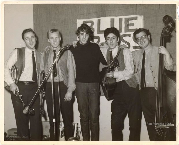 Photographie en noir et blanc de quatre hommes portant des gilets et des cravates et tenant des guitares, des mandolines et une basse sur pied, avec au centre un cinquième homme portant un pull à col roulé noir.