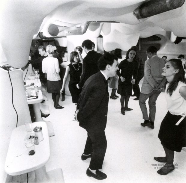 Photographie en noir et blanc d'une douzaine de personnes dansant dans une salle très éclairée, aux murs et au plafond blancs et moulés en forme de bulbe. Des tables blanches sont disposées le long du mur à gauche de l'image.