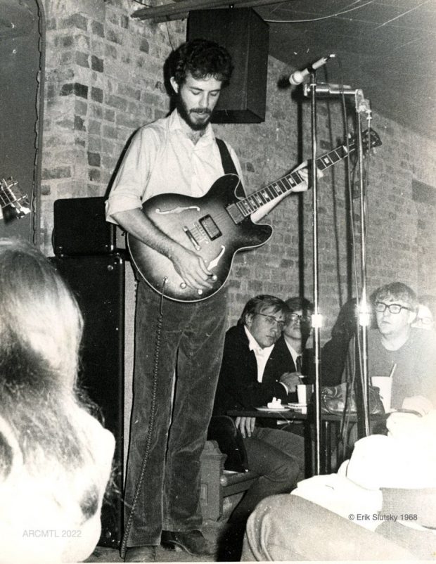 Photo en noir et blanc du chanteur Jesse Winchester, cheveux noirs mi-longs, moustache et barbe taillée, tenant une guitare dans un café avec des clients assis à côté de la scène.