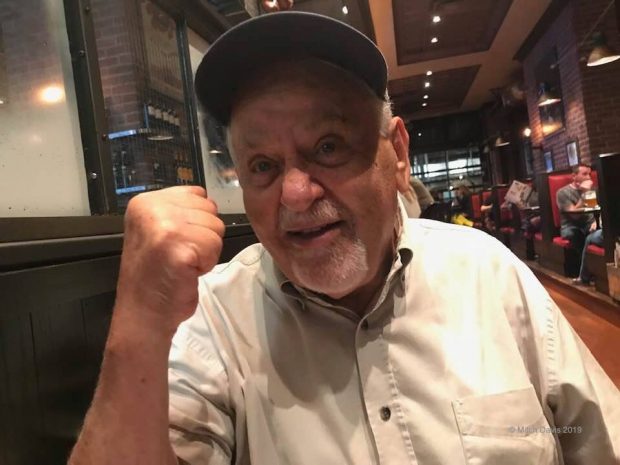 Photo en gros plan d'un Larry Kent âgé, aux cheveux blancs, assis à une table dans un restaurant, portant une casquette et le poing serré en direction de l'appareil photo.
