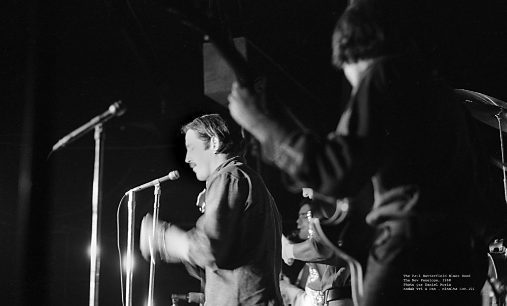 Photographie en noir et blanc de Paul Butterfield et de deux membres de son Blues Band, vus du côté de la scène lors d'un concert au New Penelope