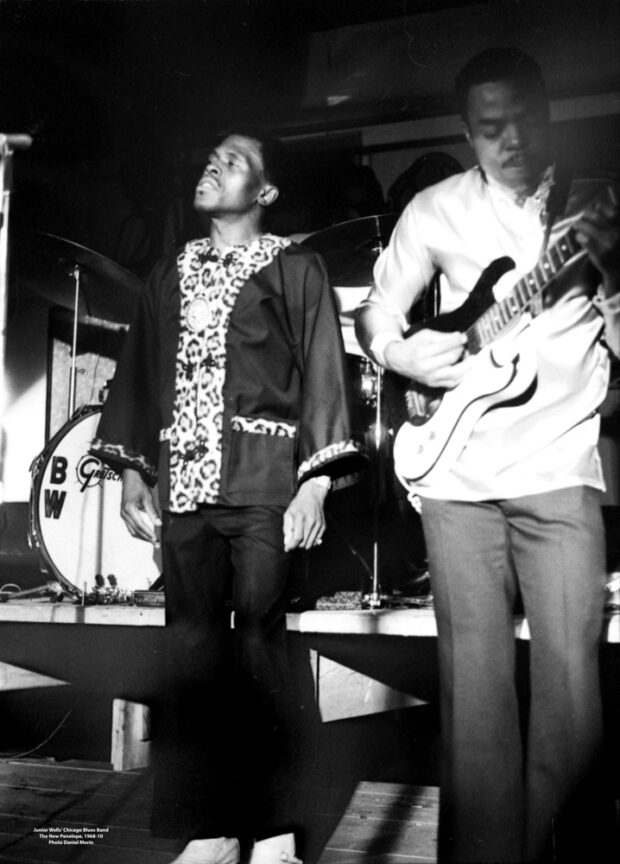 Photographie en noir et blanc représentant le chanteur de blues Junior Wells, vêtu d'un blazer à col évasé et d'un pantalon noir à fond plat, debout à côté de son guitariste, avec une partie de la grosse caisse à l'arrière-plan. Ils se produisent dans la salle de concert The New Penelope
