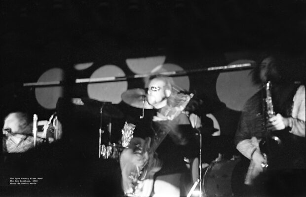 Photographie horizontale en noir et blanc montrant des motifs lumineux circulaires psychédéliques au-dessus d'un guitariste et d'un saxophoniste du Linn County Blues Band jouant en direct dans la salle de concert The New Penelope