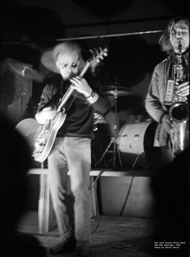 Photographie en noir et blanc montrant un guitariste et un saxophoniste du Linn County Blues Band jouant en direct devant une batterie dans la salle de concert The New Penelope
