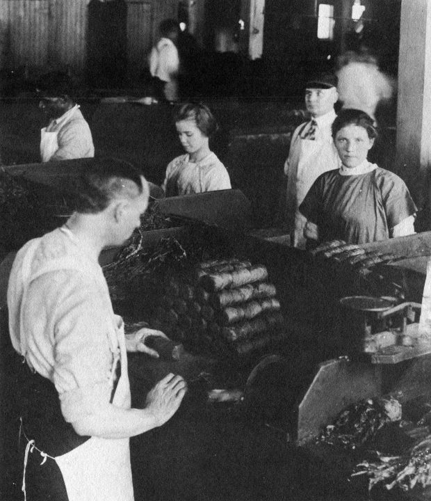 Jeunes femmes et enfants au travail à l’atelier de la manufacture de tabac Tuckett and Billings. Les femmes sont vêtues d’uniformes caractéristiques du XIXe siècle, et plusieurs d’entre elles portent également de longs tabliers de travail. On peut voir des rouleaux de tabac devant les ouvriers; une jeune femme regarde directement le photographe.