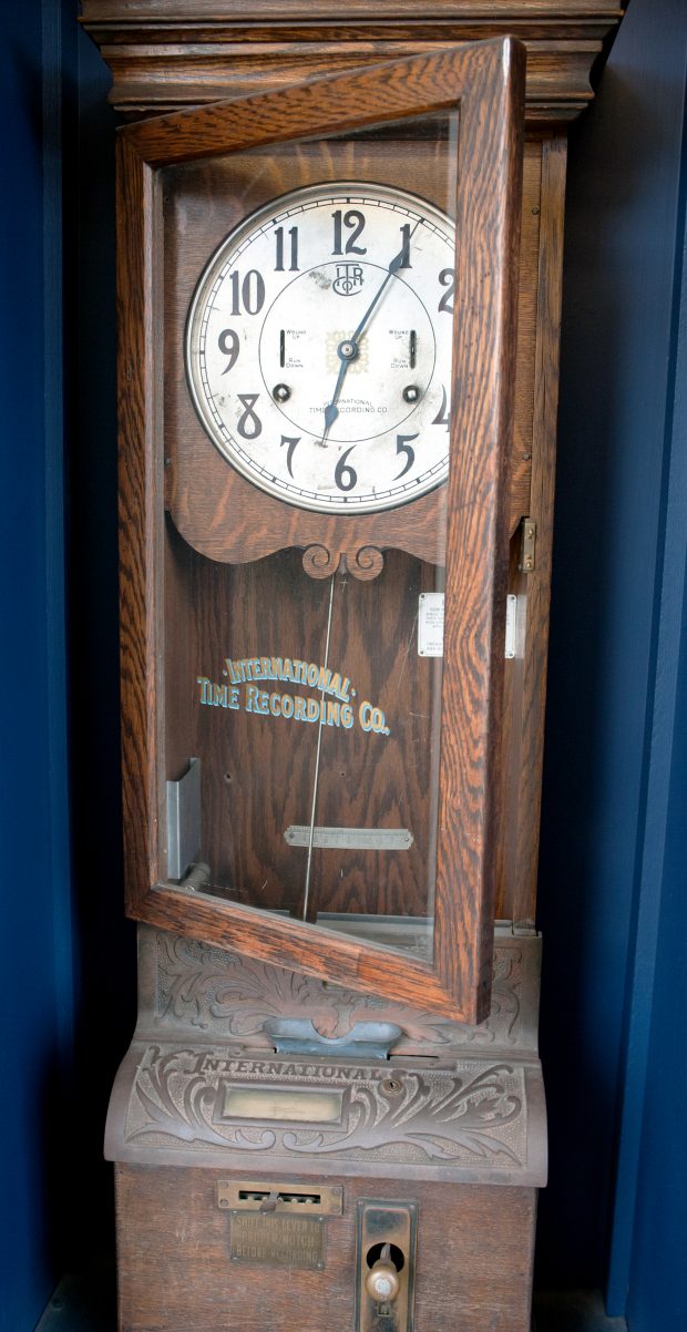 Horloge murale en bois dotée de fentes pour billets et d’un levier horodateur. Le levier doit être décalé pour inscrire l’heure sur le bordereau, puis remis en position verticale.