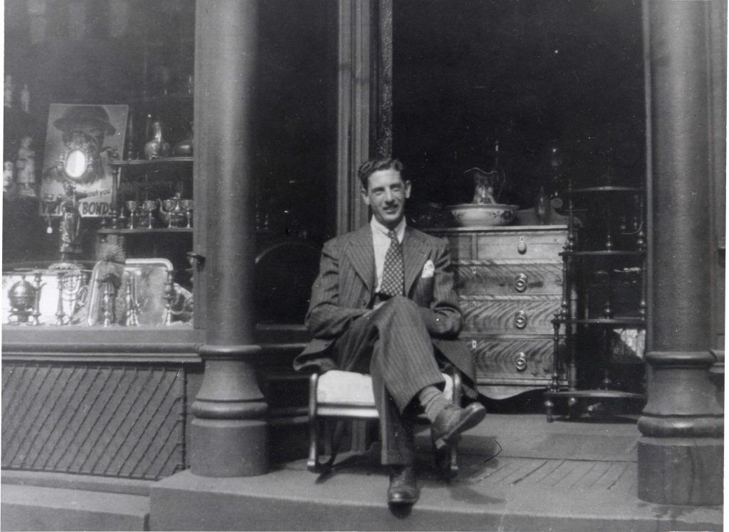 Un homme en costume est assis sur une chaise à bascule à l’entrée du magasin avec une commode et une table en bois en arrière-plan. Une vitrine à gauche est remplie d’objets.