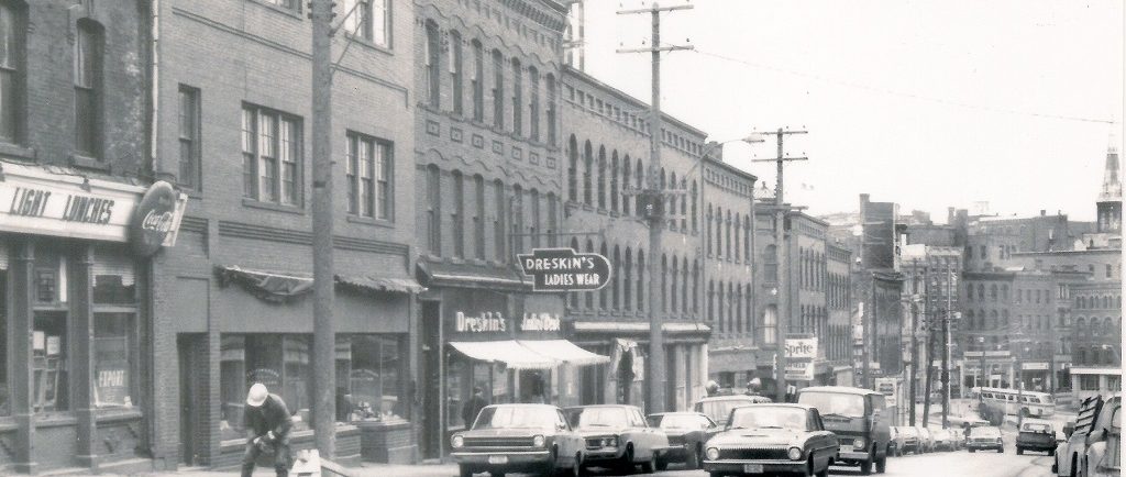 Paysage urbain avec des immeubles en briques abritant des commerces au rez-de-chaussée et des voitures typiques de la fin des années 1960 garées dans la rue. 