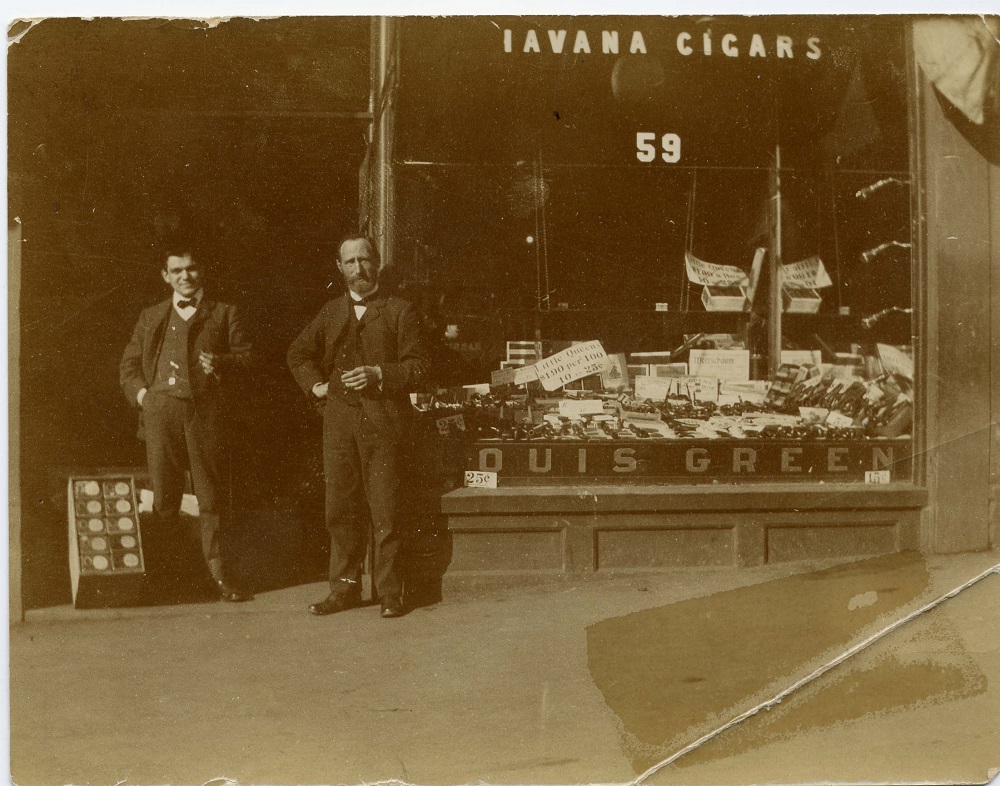 Devanture dotée d’une grande vitrine. En haut, on peut lire « Havana Cigars » et en bas, « Louis Green ». La vitrine expose des cigares et des produits de tabac. Deux hommes se tiennent debout dans l’entrée.