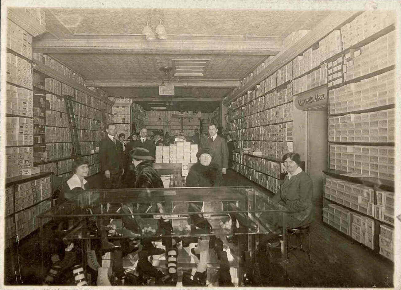 Intérieur du magasin de chaussures avec des boîtes de chaussures sur des étagères le long des murs de chaque côté. Une table en verre trône au centre, entourée de vendeuses et de clientes. Des hommes se tiennent à l’arrière-plan.