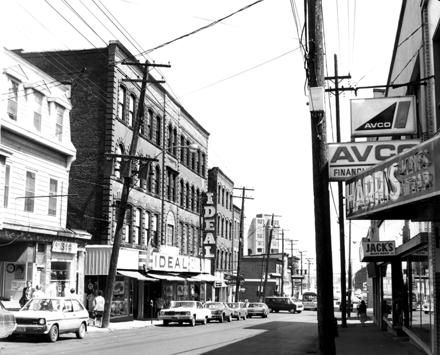 Vue d’une rue bordée d’immeubles de deux et trois étages avec des magasins au niveau de la rue et des enseignes. On distingue notamment celles de Bill’s Cigarette Centre, Ideal, Jack’s Men’s Shop et Harry’s Men’s Wear.