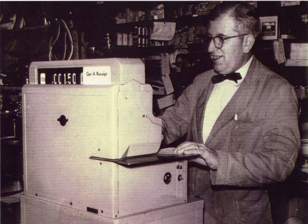 Un homme portant des lunettes à monture ronde et un tablier de magasin se tient devant une grande caisse enregistreuse. 