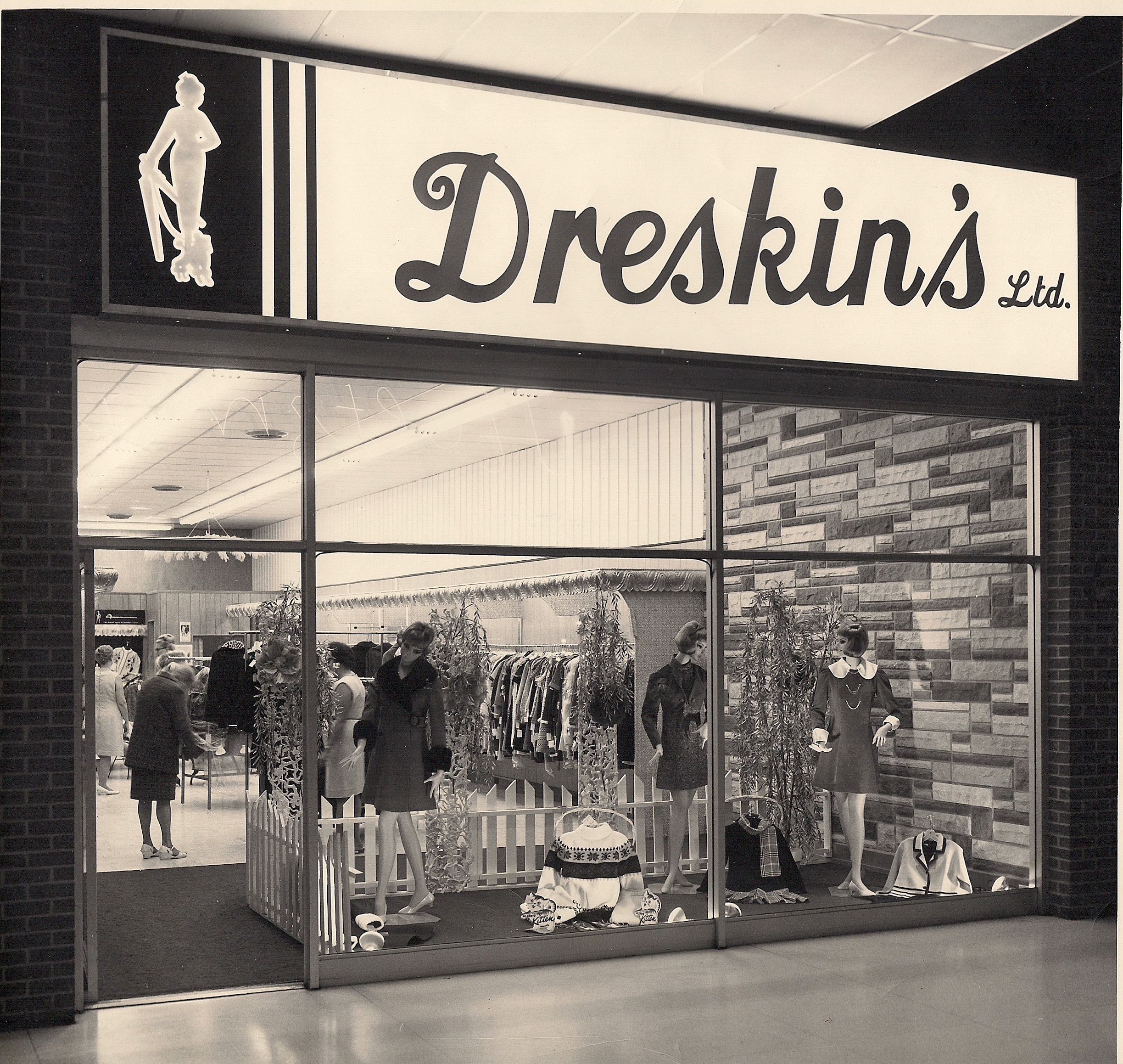 Vitrine de Dreskin’s Ltd. Trois mannequins en manteaux courts sont disposés dans la vitrine avec des présentoirs de vêtements, devant des clients qui se trouvent à l’intérieur.
