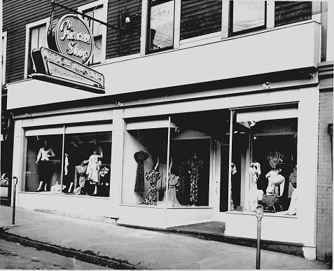 Bâtiment en bois avec trois grandes vitrines où l’on voit des mannequins en robe. L’enseigne de Princess Dress Shop est accrochée à une ferrure au-dessus de la porte.