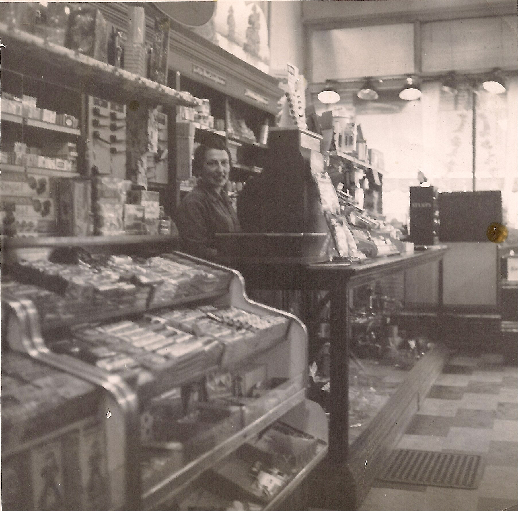Intérieur de magasin de quartier avec un comptoir rempli de snacks à gauche. Une jeune femme se tient derrière une grande caisse enregistreuse.
