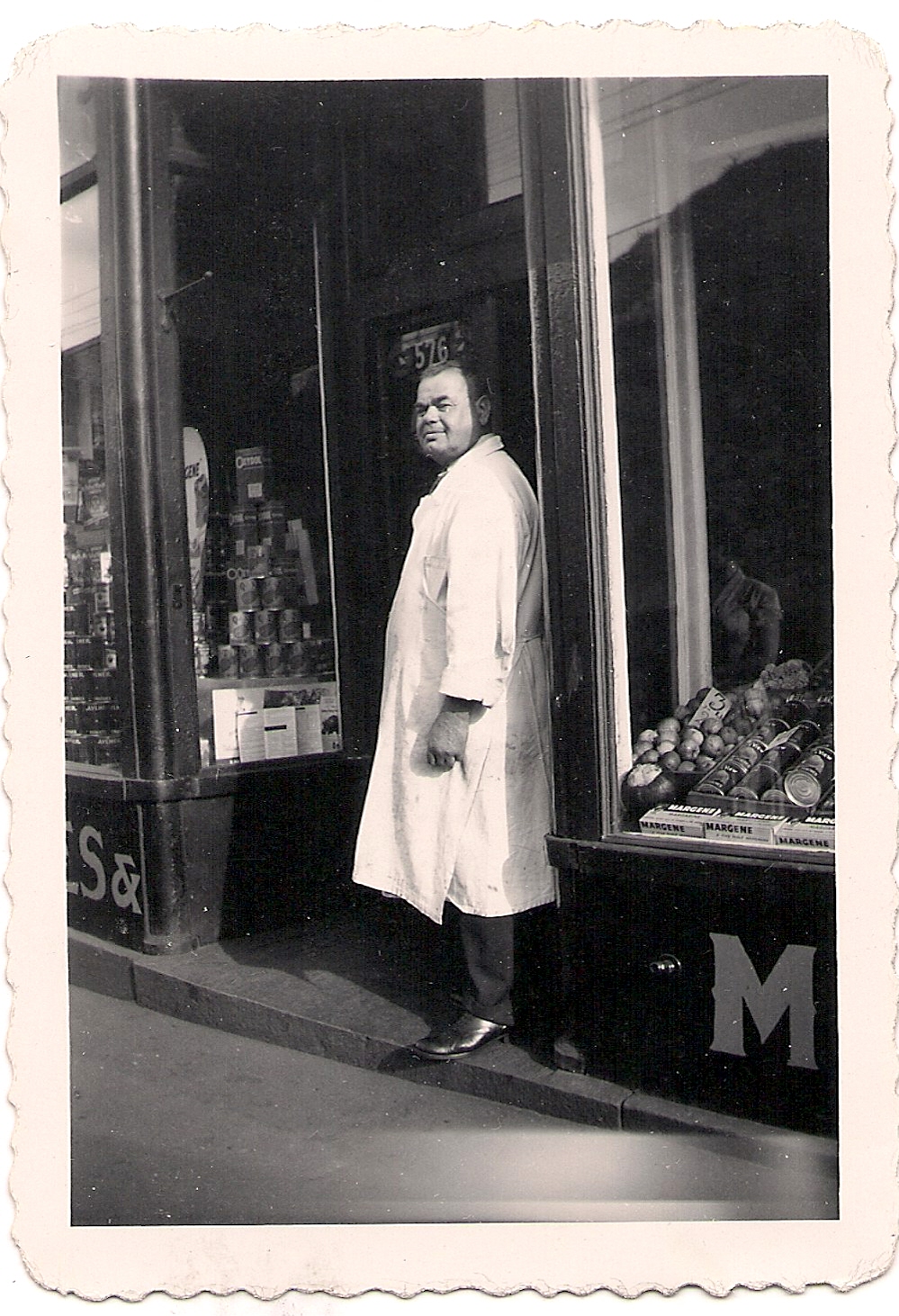 Un homme portant un long manteau blanc se tient à l’entrée d’une boucherie où l’on voit une pile de conserves en vitrine.
