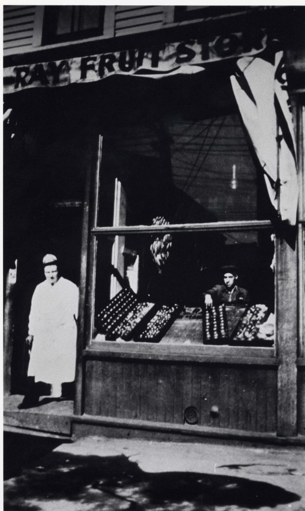 Un homme vêtu d’une longue blouse de travail se tient dans l’embrasure d’une porte à côté d’une vitrine de produits. L’inscription Sun Ray Fruit Store figure sur l’enseigne au-dessus de la fenêtre.