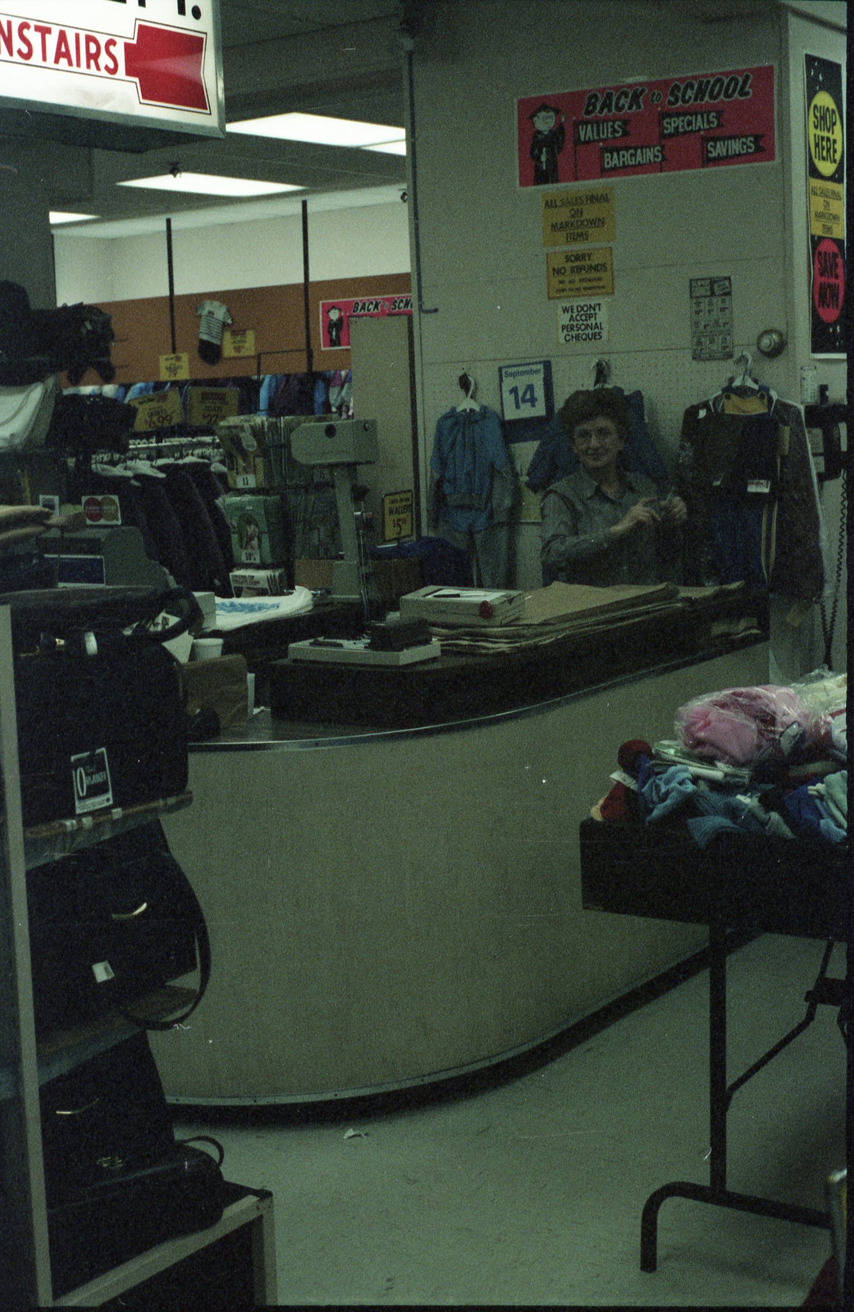 Une femme se tient derrière un comptoir incurvé, près de stands de sacs à main et de vêtements au premier plan.