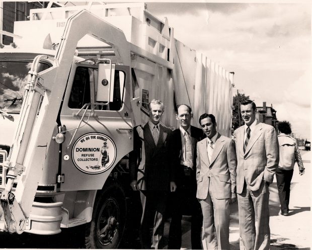 Quatre hommes se tiennent debout à côté d’un grand camion muni d’un bras avant pour vider les bennes à ordures. Sur la porte du conducteur, on voit l’inscription « We Go For Garbage - Dominion Refuse Collectors » avec un dessin représentant un chat poussant un balai.