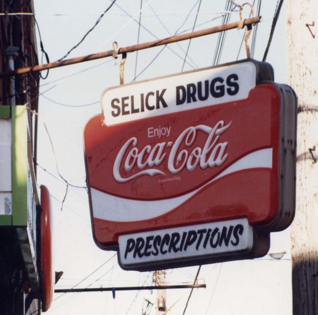 Enseigne accrochée à un support métallique sur le côté d’un bâtiment, où on lit « Selick’s Drugs » et « Prescriptions » avec un grand logo Coca-Cola entre les deux.