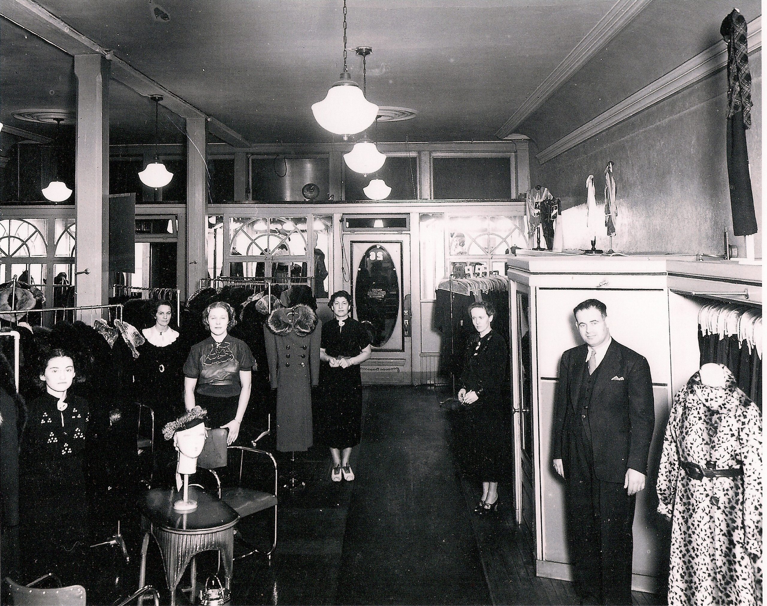 Quatre vendeuses et un homme à l’intérieur d’un magasin de vêtements pour femmes. Des robes et des manteaux sont disposés sur des présentoirs à gauche.
