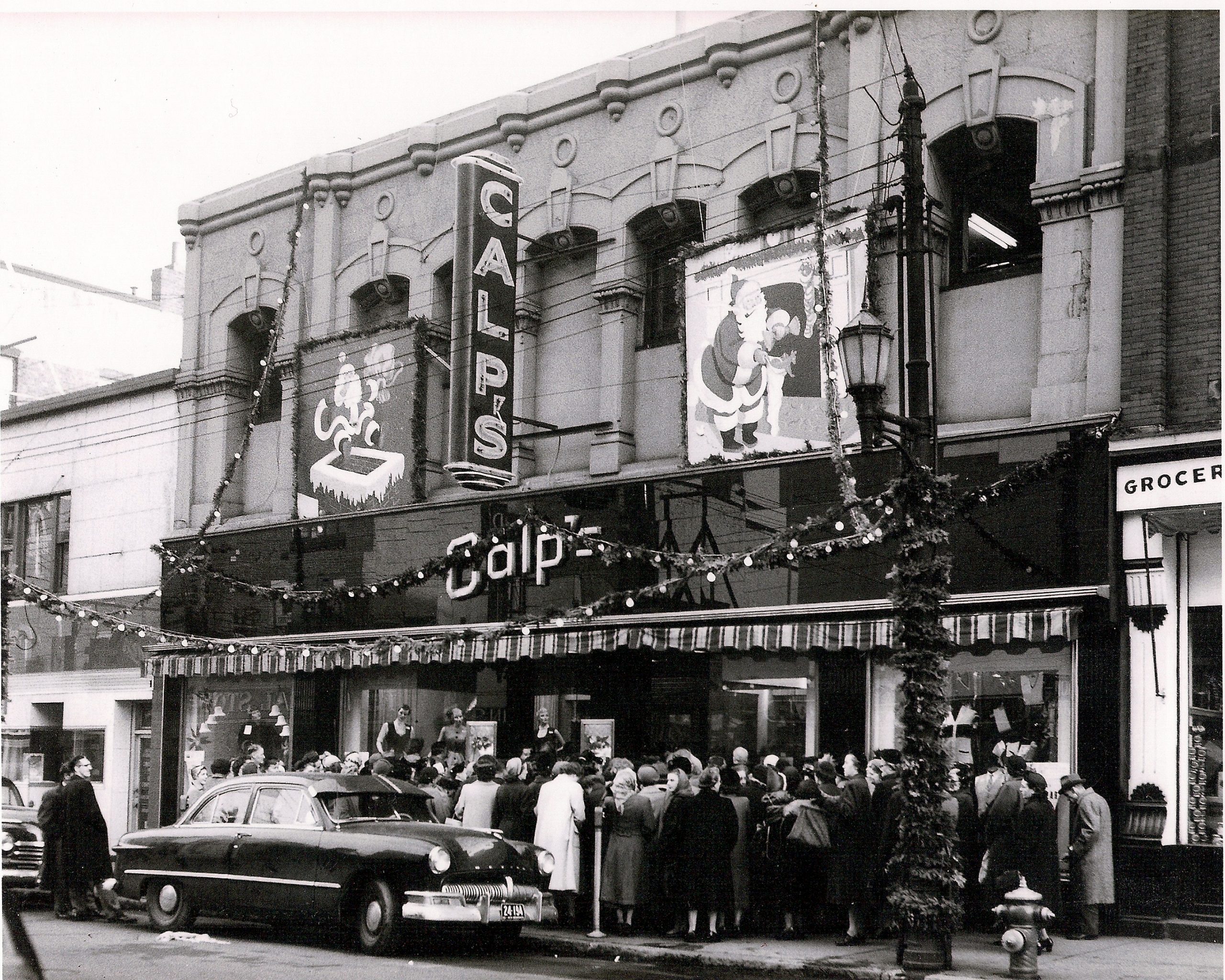 Vue extérieure du magasin Calp’s avec de grands panneaux au dessus de la porte, des décorations de Noël et une grande file de clients attendant d’entrer.