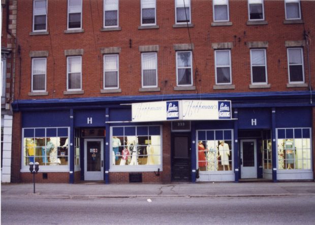Extérieur d’un bâtiment en briques avec un magasin au niveau de la rue. Le magasin a quatre vitrines et une enseigne Hoffman’s centrée au-dessus des fenêtres.
