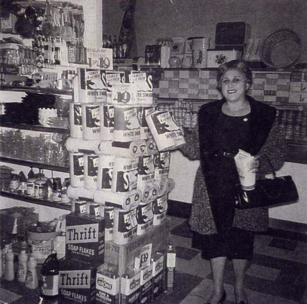 Femme dans une épicerie tenant un carton et un paquet. Les étagères à sa gauche sont remplies de marchandises.