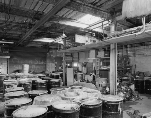 Photographie en noir et blanc représentant une salle d’entreposage industriel. C’est une grande pièce en désordre clairement laissée à l’abandon. Elle est remplie de barils de métal au contenu inconnu. 