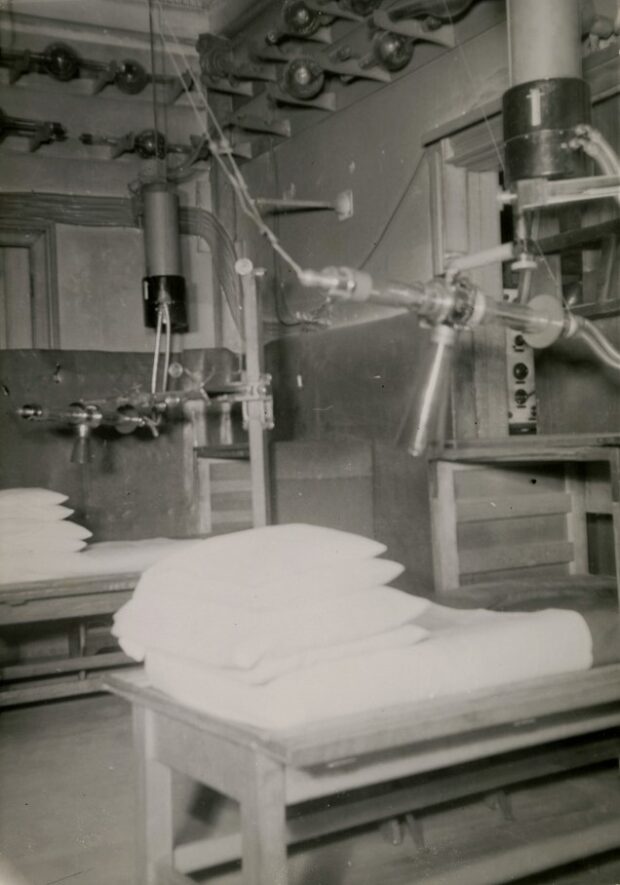 Photographie en noir et blanc représentant le coin d’une pièce remplie de machine et de filage. Deux appareils composés de plusieurs tubes reliés ensembles suspendus au plafond et attachés aux murs sont installés par dessus deux tables munies de matelas et d’oreillers. 