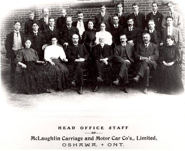 Une photographie en noir et blanc de plusieurs dizaines d’employés et employées de bureau en tenue formelle des années 1900 qui posent devant un mur de briques. La légende indique « PERSONNEL DU SIÈGE DE LA McLaughlin Carriage and Motor Car Co., Limited, OSHAWA, ONTARIO »